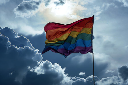 Rainbow flag waving in the wind against a blue sky with cloudsの素材