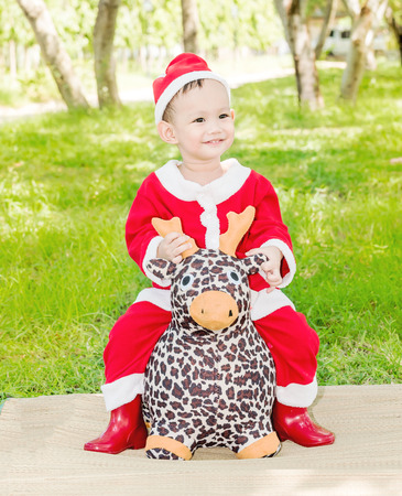 Asian baby boy in santa claus suit is playing with reindeer doll at outdoor parkの写真素材