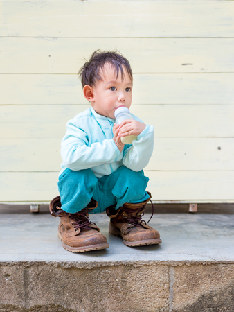 Thai Baby boy try to  wearing father's boots while drinking milk from bottle.の写真素材