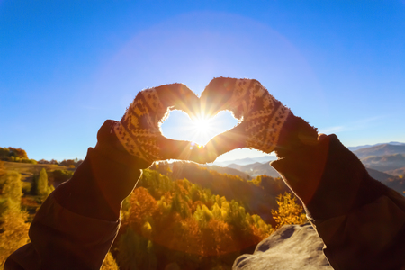 Female hands in gloves is making a heart where the sun rays shine with the background of gray stone, high mountains and golden forest in autumn day.の写真素材