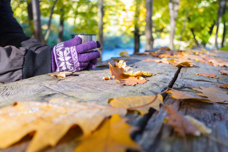 Hands in gloves hold the cap of coffee that is standing on the wooden table with yellow leaves covered by hoar frost in autumn morning.の写真素材
