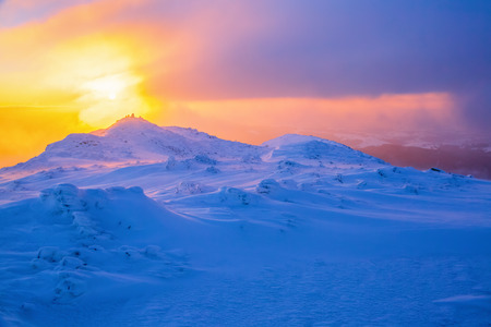 Sunrise enlightens sky, mountain and trees standing in snowdrifts covered by frozen snow with yellow shine.の写真素材