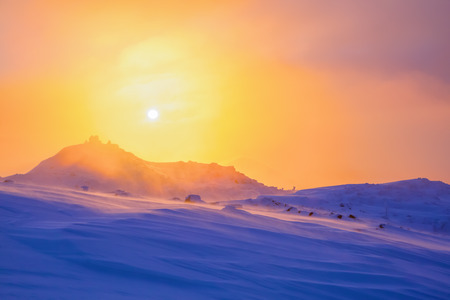 Sunrise enlightens sky, mountain and trees standing in snowdrifts covered by frozen snow with yellow shine.の写真素材