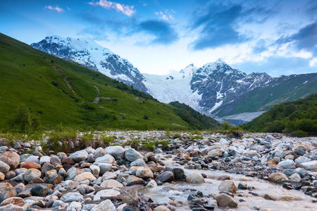From the river shore, covered with stones, opens view on fantastic glacier and steep rocky mountains with green meadows, which are covered with snow.Happy lifestyle. Beautiful universe.の写真素材
