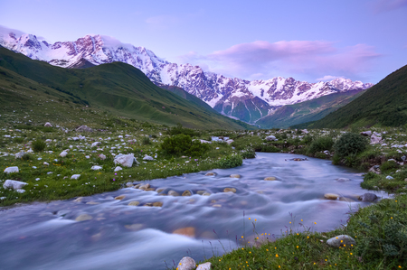 From the river shore, covered with stones, opens view on fantastic glacier and steep rocky mountains with green meadows, which are covered with snow.Happy lifestyle. Beautiful universe.の写真素材