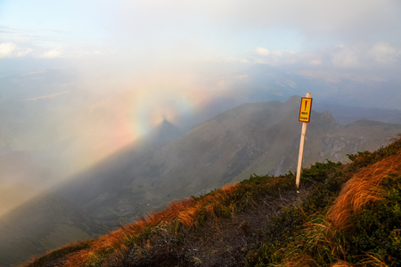 From the lawn with orange grass opens a panorama of high mountains, blue sky with clouds and a Brocken Spectre in the fog on an autumn day.の写真素材