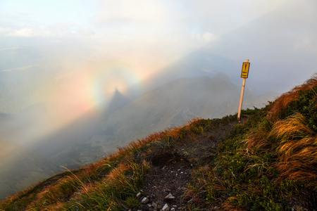 From the lawn with orange grass opens a panorama of high mountains, blue sky with clouds and a Brocken Spectre in the fog on an autumn day.の写真素材
