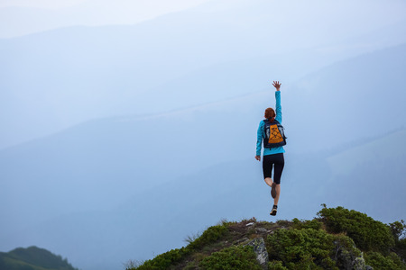 Red hair girl with the back sack jumps on one leg from happiness among the high mountains at the edge of the cliff.の写真素材