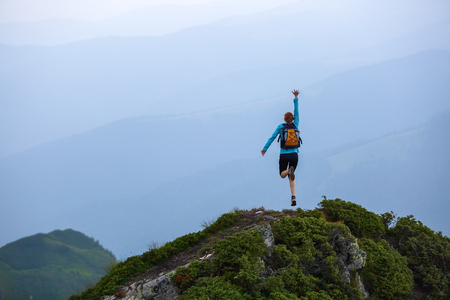 Red hair girl with the back sack jumps on one leg from happiness among the high mountains at the edge of the cliff. Location the Carpathian Mountains, Marmarosy, Ukraine.の写真素材