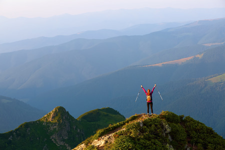 The tourist girl with the tracking sticks and the back sack at the edge of the cliff. The landscape with the high mountains. Endless vastness on the horizon. Magnificent evening summer landscape.の写真素材