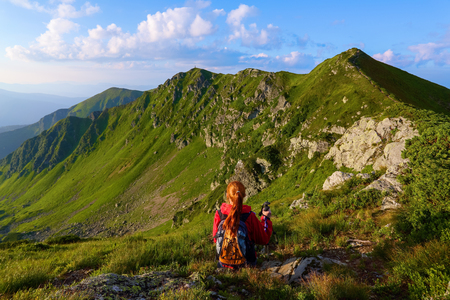 The view with the high rocky mountains. A young girl sits on the green grass among the flowers. Sur rays. Beautiful clouds are slowly moving in the sky. Summer scenery.の写真素材