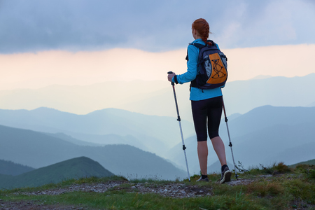 The tourist girl with back sack and tracking sticks is staying on the lawn. Beautiful cloudy sky. Sun rays fall down on the mountains. Summer scene. . Eco touristic laisure time.の写真素材