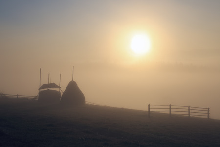 The bright disk of the sun illuminates with a warm orange light the mountain meadow where loose stacked hays built around a central pole stand, which are shrouded in a light mist.の写真素材