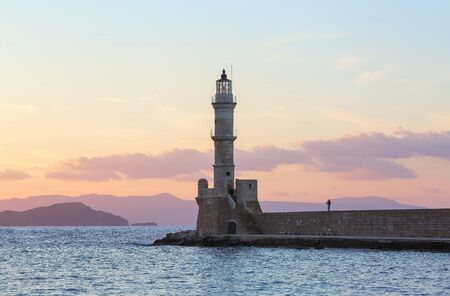High, beautiful, ancient lighthouse made of bricks. Marvelous sunset lights the sky. Touristic resort Chania, Creete island, Greece. Along the sea shore there is a wall and the road.の写真素材