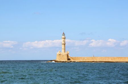 Ancient brick lighthouse. The tourists are standing on the wall. Popular place for holidays seaport Chania, Creete island, Greece. Seafront along the emerald sea with waves. Blue sky with cloudsの写真素材