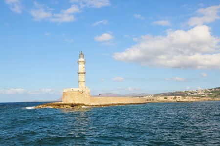 Ancient brick lighthouse. The tourists are standing on the wall. Popular place for holidays seaport Chania, Creete island, Greece. Seafront along the emerald sea with waves. Blue sky with cloudsの写真素材