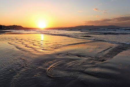 Location place Agia Marina Beach, island Crete, Greece. Sea coast spangled by rocks, the sunrise is reflecting on the wet sand. The mountains in haze on the horizon. Storm sea with high waves.の写真素材
