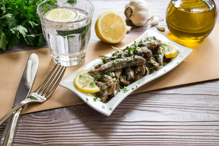 Freshly cooked sardines on a plate near fork and knife, glass of sparkling water, bunch of parsley, half a lemon, garlic, olive oil. Fresh sardines for lunch. Horizontal. Daylight.の写真素材