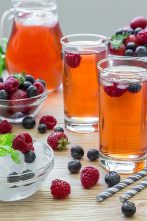 2 compotes of raspberries, strawberries, blueberries, near bowl of ice, 2 bowls with berries, mint leaves, carafe compote, straws on light background scattered berries. Berries compote. Vertical.の写真素材