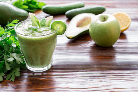 Left glass of green vegetable smoothie near ingredients celery, avocado, cucumber, apple, lemon and herbs, right empty space on wooden background. Green vegetable smoothie and ingredients. Horizontal.の写真素材