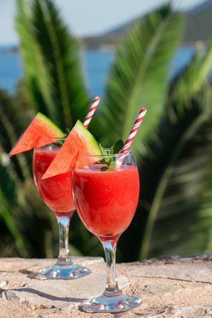 Two watermelon smoothie on the terrace overlooking the sea on a background of palm branches. Watermelon smoothie on summer holidays by the sea. Vertical. Daylight. Close.の写真素材