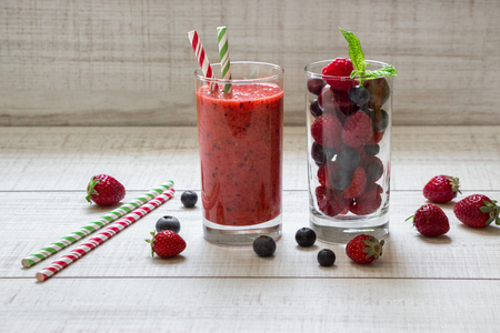 A glass of smoothie with berries and a glass of ingredients strawberries, raspberries, blueberries near scattered berries, straws on white wood background. Berry smoothie and ingredients. Horizontal.の写真素材
