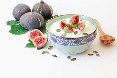 A dish of yogurt with pieces of fig fruit, near scattered sunflower seeds, laid ripe fig fruits on the fig tree leaves on the white background. Yogurt with fig fruit slices. Horizontal. Daylight.の写真素材
