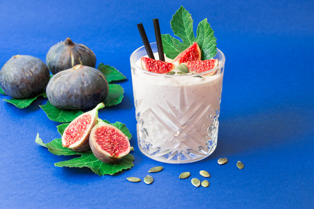 Glass of fig fruit smoothie, fig slices, straws and near scattered sunflower seeds, ripe fig fruits on the fig tree leaves on the blue background. Fig fruit smoothie glass. Horizontal. Daylight.の写真素材
