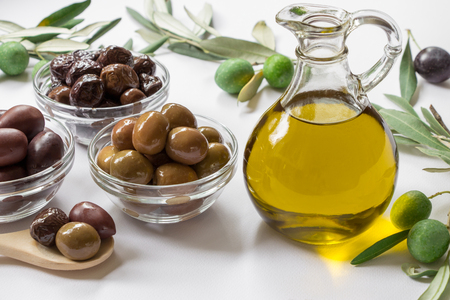Glass bottle with premium virgin olive oil near variety of olives in bowls and spoon, olive tree branches on the white background. Premium virgin olive in bottle and variety of olives. Horizontal.の写真素材
