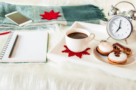 Cup of coffee, sweets, alarm clock on a tray near notebook, pencils, holiday decorations, green checkered scarf, mobile on white artificial fur background. Breakfast time New year planning concept. Horizontal.の写真素材