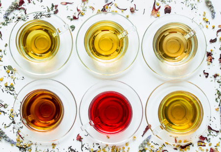 Six cups with different types of tea black, green, hibiscus, chamomile, peppermint, lavender on white background of scattered tea leafs. Six types of tea on a white background of scattered tea leafs.の写真素材