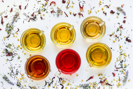 Diagonally in 2 rows 6 cups with different types of tea black, green, hibiscus, chamomile, peppermint, lavender on white background of scattered tea leafs. Six types of tea on a white diagonal lines.の写真素材