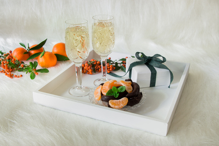 Tray with champagne in 2 crystal glasses, tangerines slices, chocolate and gift on background of rowan twigs, tangerines all on an artificial white fur. Champagne, chocolate, tangerines. Horizontal.の写真素材