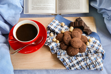 Breakfast served in bed - cup of coffee, chocolate cookies, chocolate, hazelnuts on a wooden board on background of light blue bed linens, open book. Festive breakfast in bed concept.の写真素材