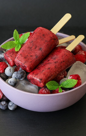 Homemade raspberries blueberries ice cream popsicles decorated berries and green mint leaves in bowl with ice cubes on dark background. Vertical. Close-up.の写真素材