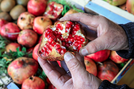 Half of the ripe pomegranate fruit in the hands of the seller in the greek vegetable shop on the background of pomegranate fruits. Horizontal. Outdoor. Close-up.の写真素材