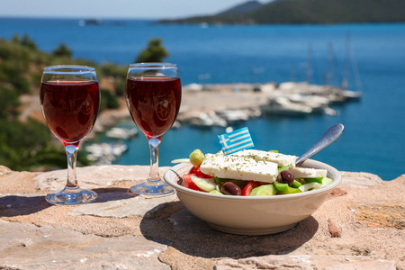 Two glasses of red wine and bowl of greek salad with greek flag on by the sea view, summer greek holidays concept.の写真素材