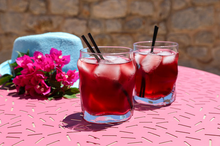 Two glasses of summer red cocktail with ice next to a blue hat and a sprig of Bougainvillea flowers on a pink table, summer Mediterranean holidays concept.の写真素材