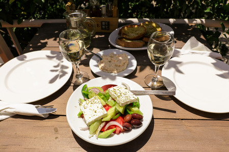 Village greek salad on white plate near glasses of white wine, hot pepper cheese dip dish, plate of bread on wooden table in greek tavern. Horizontal. Close-up. Daylight. View from above.の写真素材