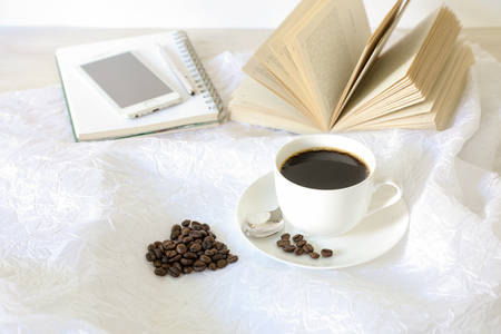 A cup of coffee, coffee beans laid out in the shape of a heart, behind an open book, notebook, mobile phone, pen on a white background.の写真素材