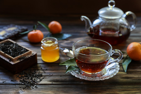 Cup of black tea, tea leaves, teapot, honey and tangerines on the wooden background. Horizontal. Daylight. Close-up.の写真素材