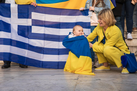 SYNTAGMA SQUARE, ATHENS, GREECE - April 3rd, 2022: Toddler little patriot wrapped in Ukrainian flag on the background of Greek flag at a peaceful protest against the WAR in UKRAINE. Horizontal.のeditorial素材