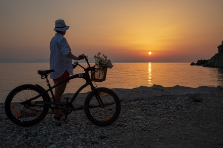 A young woman with a bicycle enjoys the sunset by the sea.の写真素材