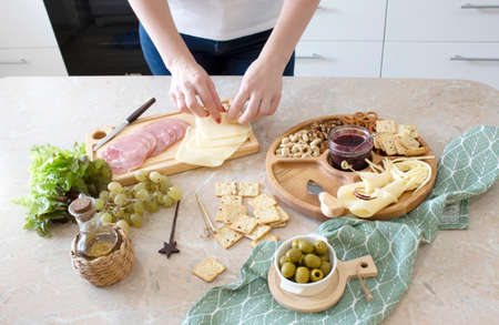 womans hands cutting cheese on wooden cutting board on wooden table with grapes and dried fruits, nuts, pumpkin seeds on wooden cheese plate on table with strip tablecloth.の写真素材
