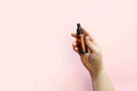 Woman hands holding facial essential oil or serum packaging on pink background. Beauty cosmetic...