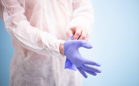 Male doctor in white virus protective suit puts on medical rubber gloves for work in blue.