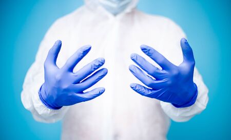 A doctor is a man in a white virus-protected suit and blue medical rubber gloves that holds out in front of his hand showing them.の写真素材