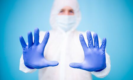 A male doctor in a white virus-protected suit and blue medical rubber gloves with a mask on his...