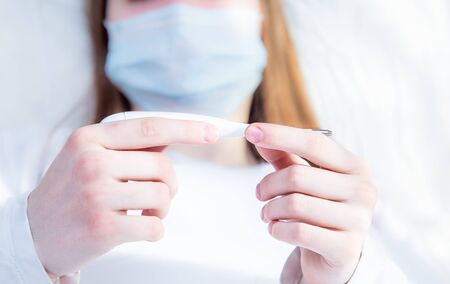 A young woman lies in bed in a mask at home. In the hands of a woman is an electronic thermometer...
