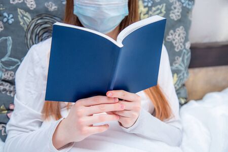 A young woman, at home on self-isolation, quarantine has the flu and lies in bed. In the hands of a girl is a book for reading in blue. medical conceptの写真素材